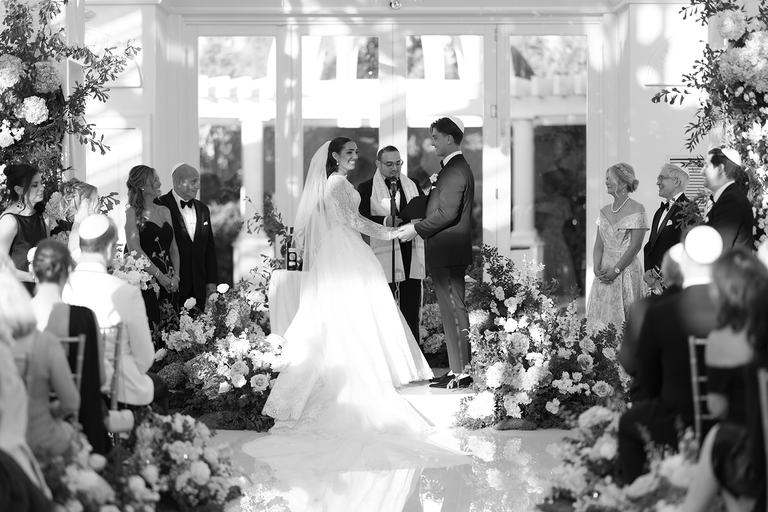 Black and white photo of a couple holding hands beneath the chuppah, surrounded by family, as Todd Bronshtein officiates