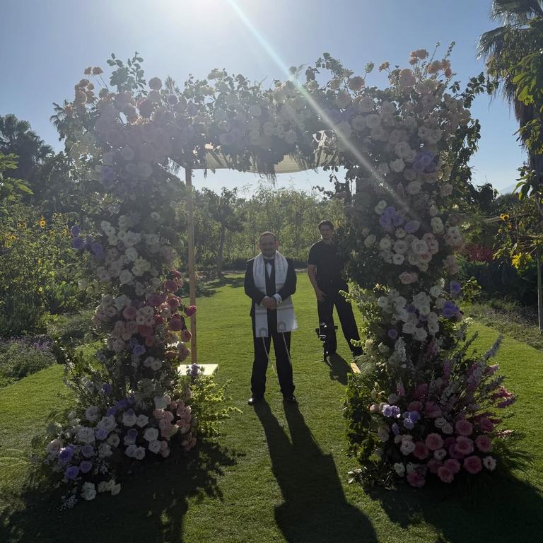 Sunlight streams through a chuppah covered in flowers in a garden before the ceremony begins
