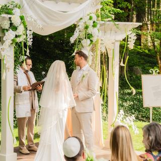 A couple beneath a white draped chuppah in a wooded garden as Todd Bronshtein officiates