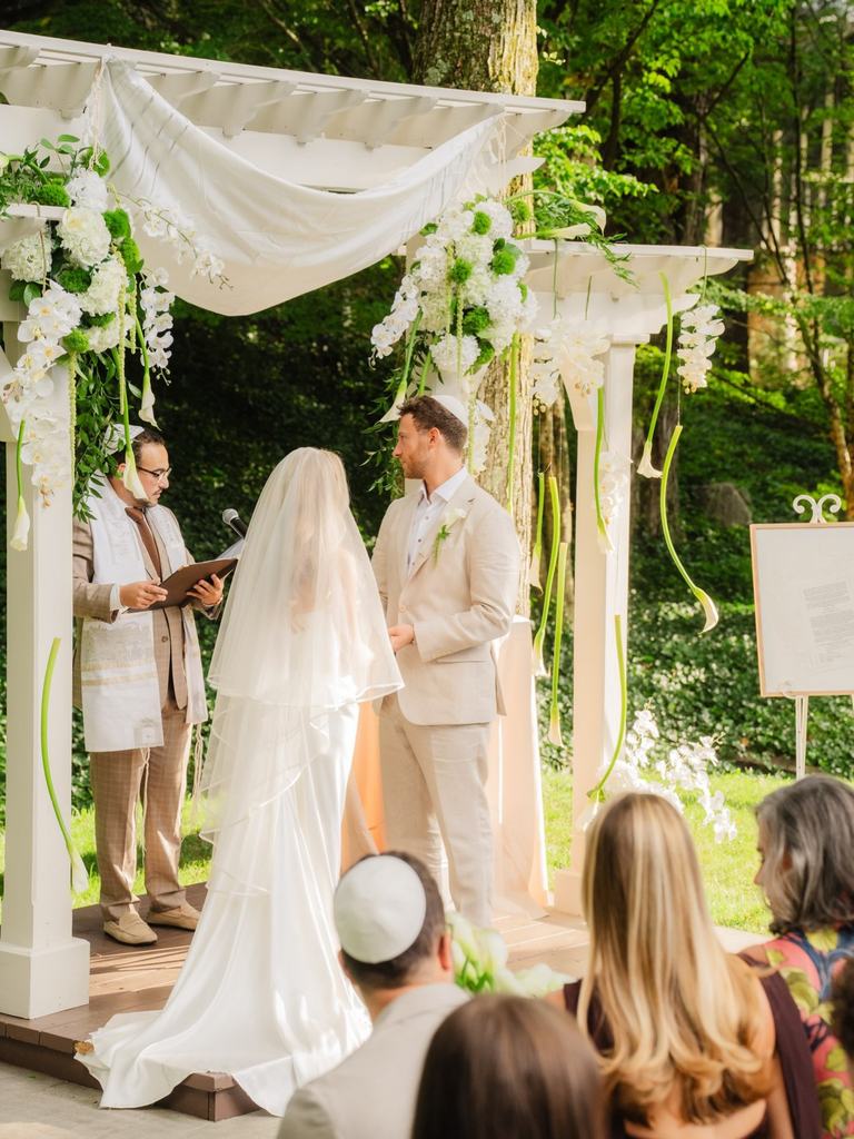 A couple stands beneath a white draped chuppah in a garden during their Jewish wedding ceremony